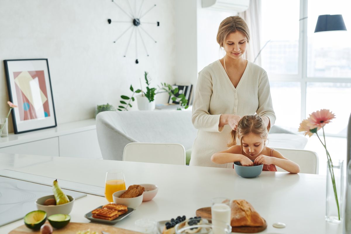 A mother braids her daughter's hair in a bright kitchen, surrounded by breakfast items. Eine Mutter flechtet ihrer Tochter in einer hellen Küche, umgeben von Frühstücksspeisen, die Haare.