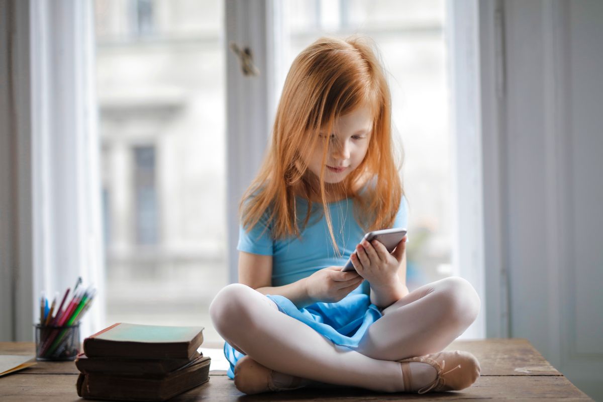 Photo by Andrea Piacquadio on Pexels Rothaariges, charmantes Schulmädchen in blauem Kleid surft auf dem Smartphone, während es zu Hause auf einem rustikalen Holztisch mit überkreuzten Beinen neben Büchern vor einem großen Fenster sitzt – Foto von Andrea Piacquadio auf Pexels
