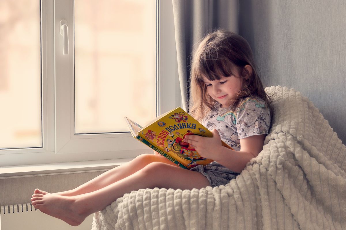 Photo by Ольга Жарикова on Pexels Ein junges Mädchen liest ein buntes Buch am Fenster und genießt die gemütliche und friedliche Freizeit drinnen. - Foto von Ольга Жарикова auf Pexels
