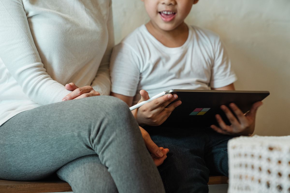 Exited crop schoolchild surfing internet on tablet while resting together with mother in domestic cloth at home during weekend Exited crop schoolchild surfing internet on tablet while resting together with mother in domestic cloth at home during weekend