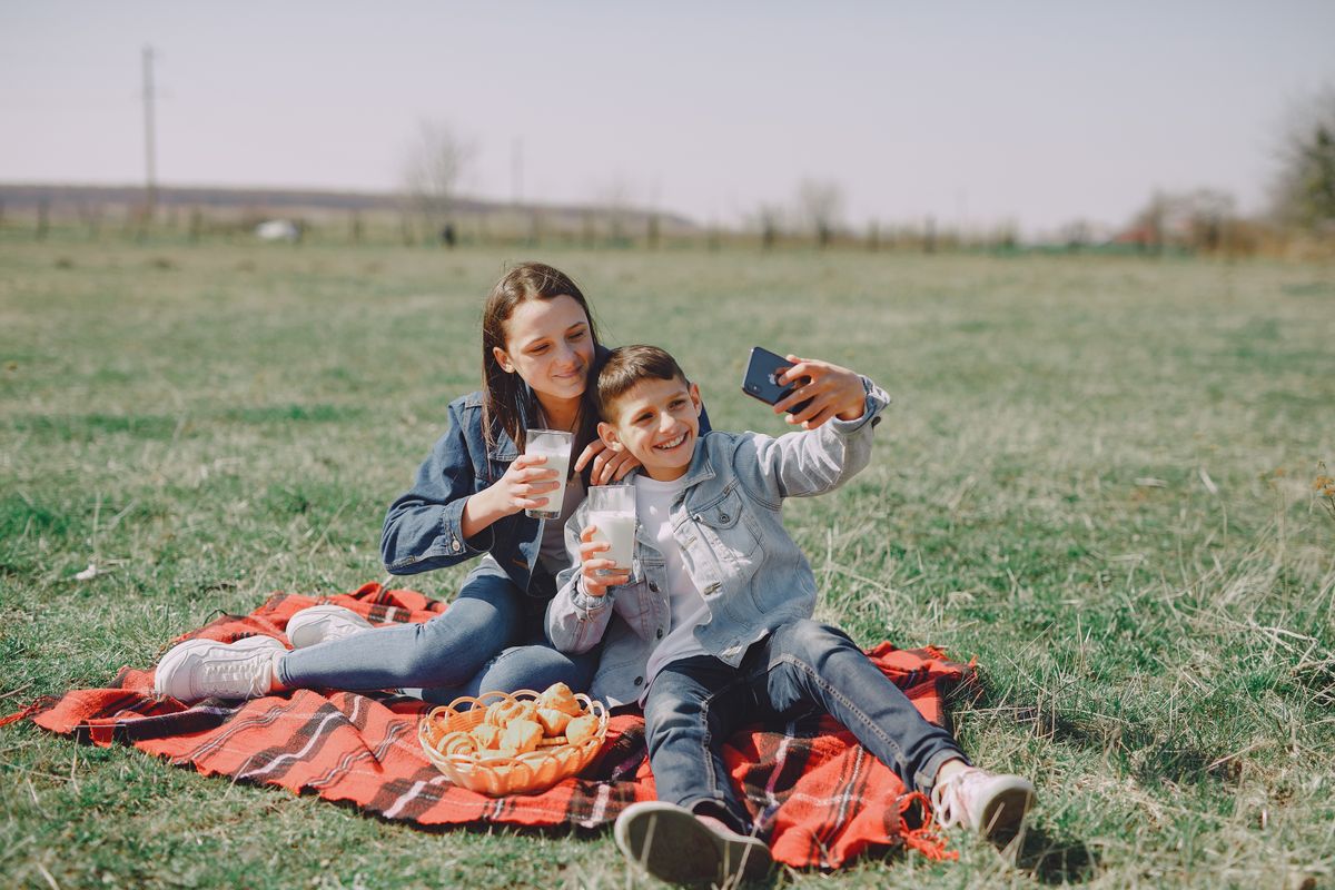 Photo by Gustavo Fring on Pexels Smiling young boy and girl in casual outfit sitting together on checkered plaid with glass of milk and buns while taking selfie on mobile phone in park - Photo by Gustavo Fring on Pexels