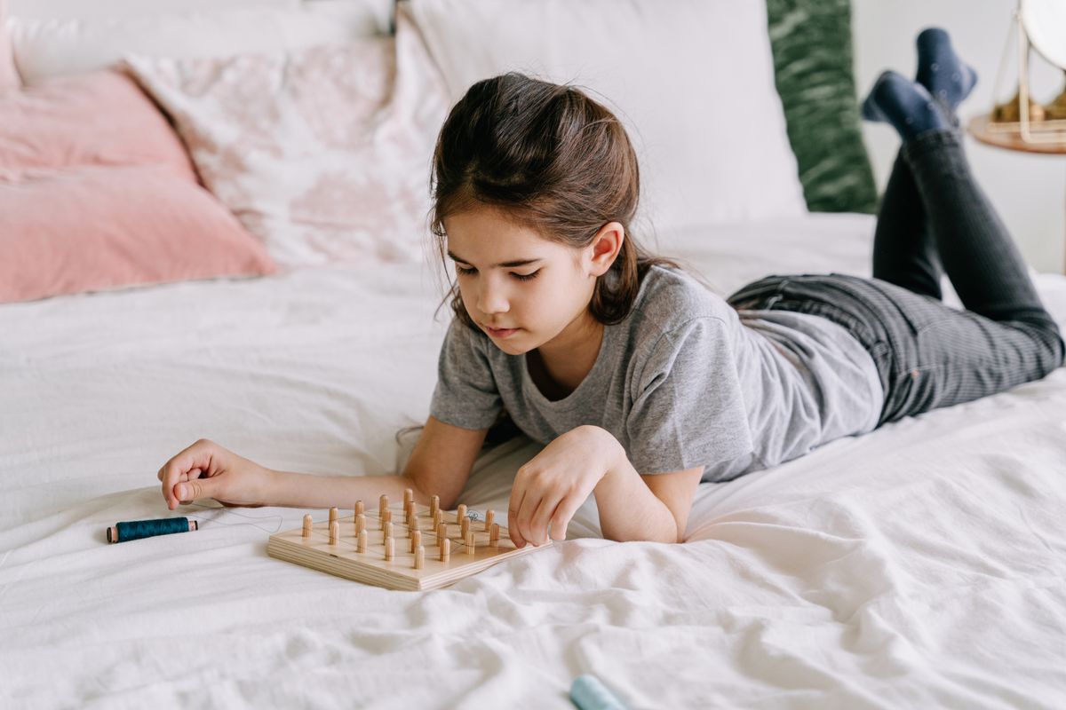 Photo by Ivan S on Pexels Ein junges Mädchen, das auf einem Bett liegt, vertieft sich in eine achtsame Geoboard-Aktivität im Innenbereich. - Foto von Ivan S auf Pexels
