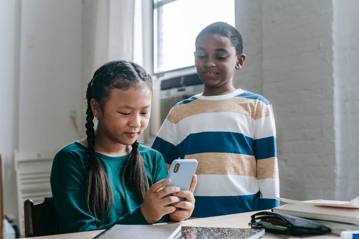 Photo by Katerina Holmes on Pexels Happy little African American boy standing near Asian classmate girl using smartphone sitting at desk during break - Photo by Katerina Holmes on Pexels
