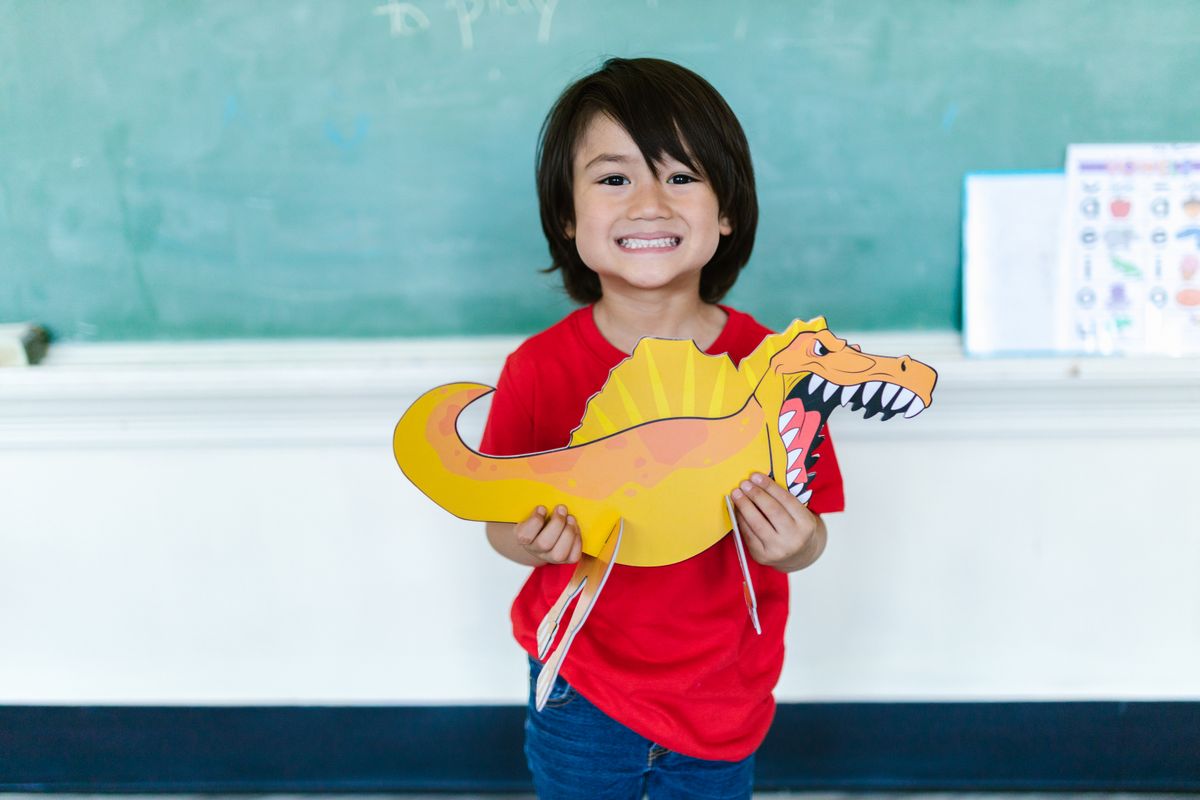 Photo by RDNE Stock project on Pexels Ein fröhliches Kind hält in einem Klassenzimmer einen bunten Dinosaurierausschnitt in der Hand. - Foto von RDNE Stock-Projekt auf Pexels