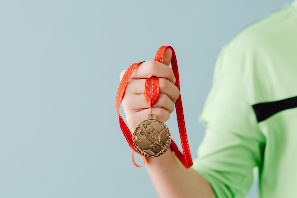 Photo by www.kaboompics.com on Pexels Nahaufnahme eines Teenagers, der eine Fußballmedaille an einem roten Band vor blauem Hintergrund hält. - Foto von www.kaboompics.com auf Pexels